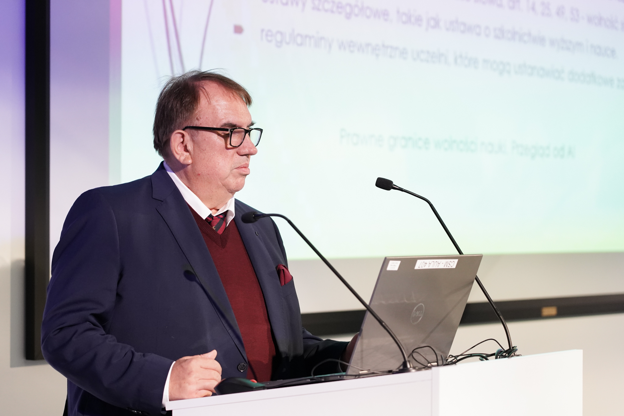 Elegancko ubrany mężczyzna w średnim wieku przed m&oacute;wnicą z laptopem i dwoma mikrofonami, w okularach, z tyłu wyświetlana część prezentacji. Mężczyzna patrzy w d&oacute;ł.